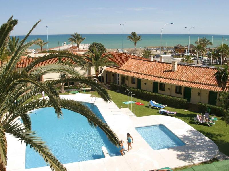Schwimmbad vor orangefarbenem Bungalow mit Palmen, dahinter blauer Himmel und Meer am Horizont.