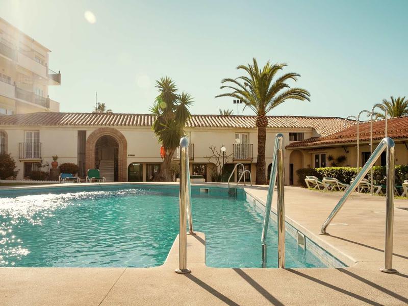 Helles Schwimmbecken mit Treppen, umgeben von Palmen und umliegenden Gebäuden bei sonnigem Wetter.