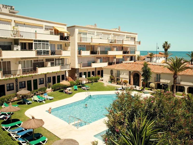 Schwimmbad mit Sonnenliegen, umgeben von weißen Wohngebäuden und Palmen unter blauem Himmel.