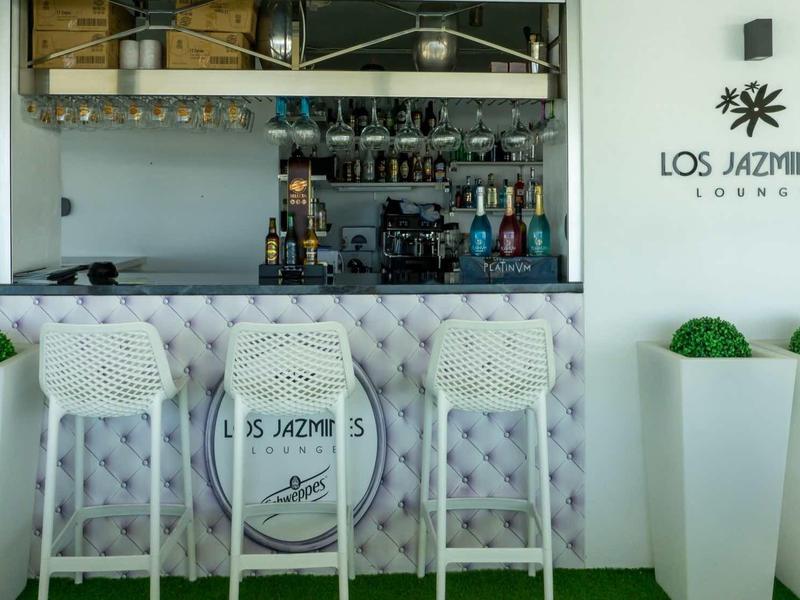 Moderner weißer Barbereich mit Hockern, grünen Pflanzen und Logo "Los Jazmines Lounge".