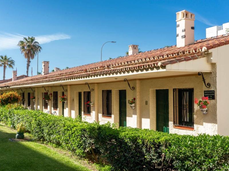 Langgestrecktes, helles Gebäude mit grünlichen Türen und Fenstern, grüner Hecke und Palmen im Hintergrund.