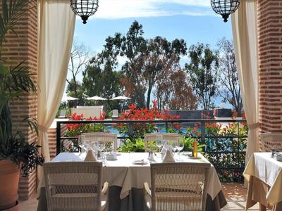 Terrasse mit gedeckten Tischen, grünen Pflanzen und Blick auf Bäume und Himmel hinter Metallgeländer.