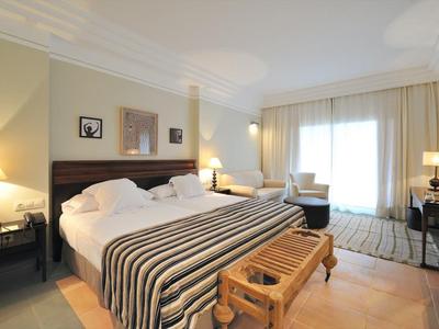 Helles Schlafzimmer mit Doppelbett, gestreifter Decke, Holzbank, Teppich und großen Fenstern.