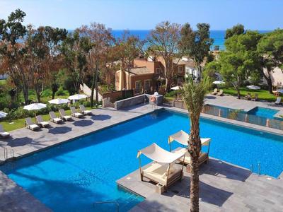 Großer blauer Pool mit Liegen und grünen Bäumen, Gebäude und Meer im Hintergrund.