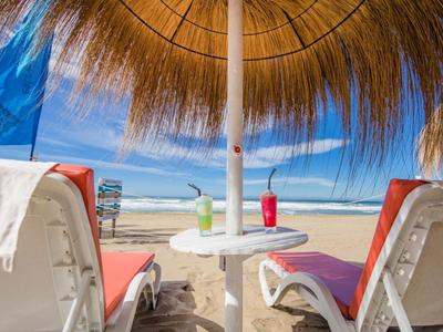 Dwa leżaki z ręcznikami i napojami pod słomianym parasolem na plaży z widokiem na morze.