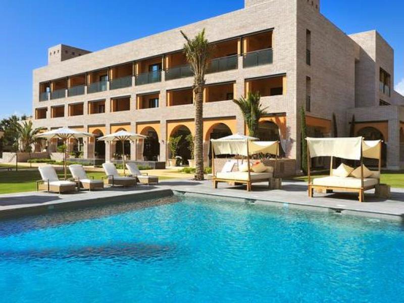 Modernes, dreistöckiges Gebäude mit großen Fenstern, Pool und Palmen unter klarem blauem Himmel.