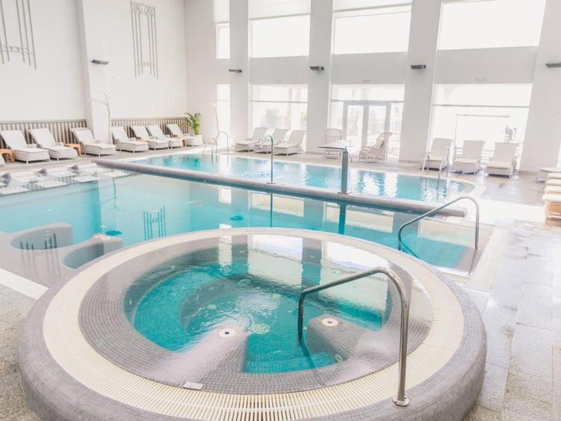 Heller Wellnessbereich mit rundem Whirlpool und großem Schwimmbecken, umgeben von Liegestühlen.