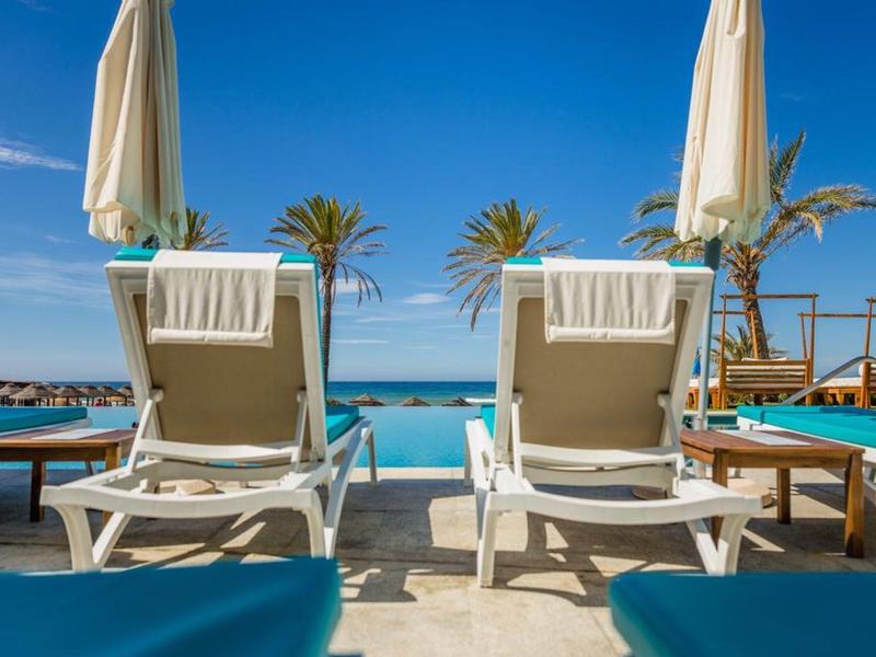 Zwei weiße Liegestühle mit Handtüchern vor blauem Meer, Palmen und blauem Himmel.