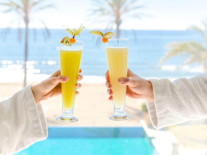 Zwei Menschen stoßen mit bunten Fruchtsäften am Pool mit Meerblick an.