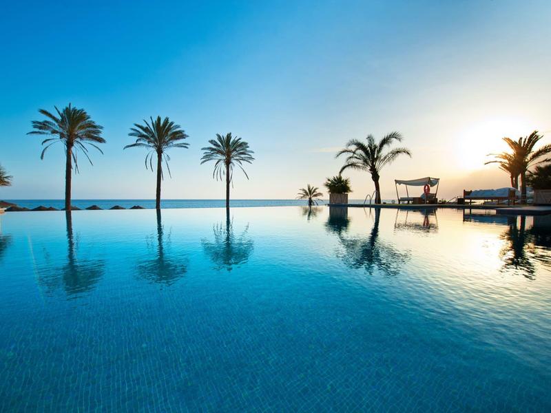Ein ruhiger Pool mit spiegelglattem Wasser und Palmen vor einem klaren blauen Himmel bei Sonnenuntergang.