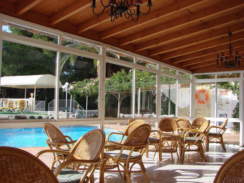 Innenraum mit Holzdecke, vielen Rattanstühlen und Tischen neben einem Pool hinter Glaswänden.