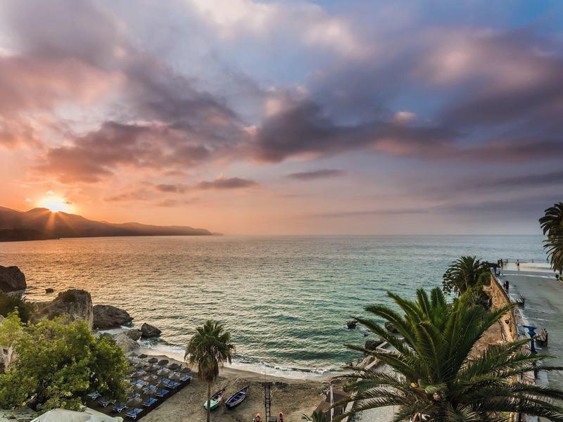 Sonnenuntergang über ruhigem Meer mit Palmen, Strand und Pier im Vordergrund.