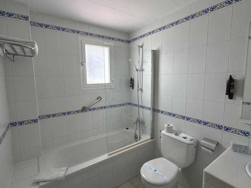 Kleines Bad mit weißer Badewanne, WC, Waschbecken und weißen Wandfliesen mit blauer Bordüre.