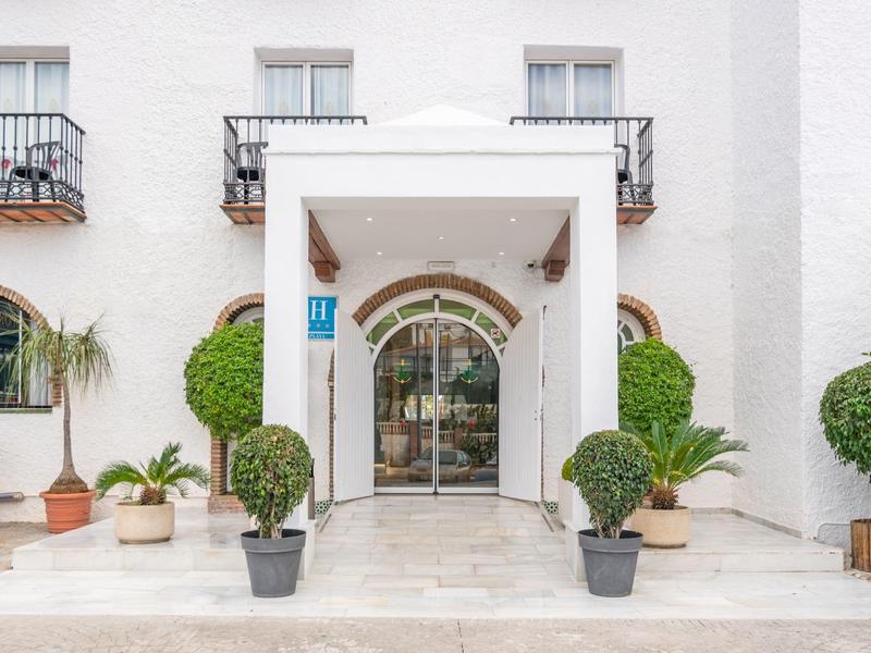 Eingangsbereich eines weißen Gebäudes mit Glas-Eingangstüren, grünen Pflanzen und Fensterbalkonen.