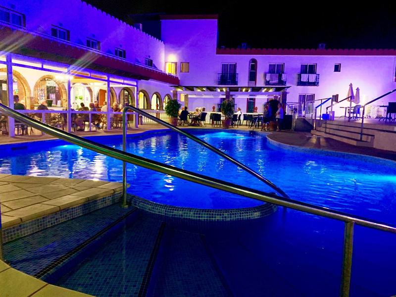 Beleuchteter Außenpool bei Nacht mit Handlauf, umgeben von einem Gebäude mit Arkaden und Terrassen.
