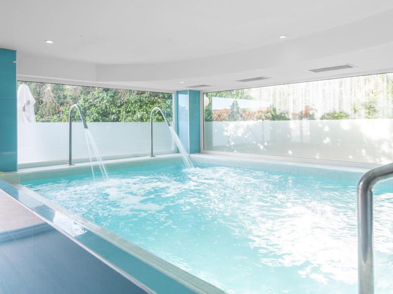 Helles Hallenbad mit blauen Säulen, Wasserstrahlen und großen Fenstern mit Blick ins Grüne.