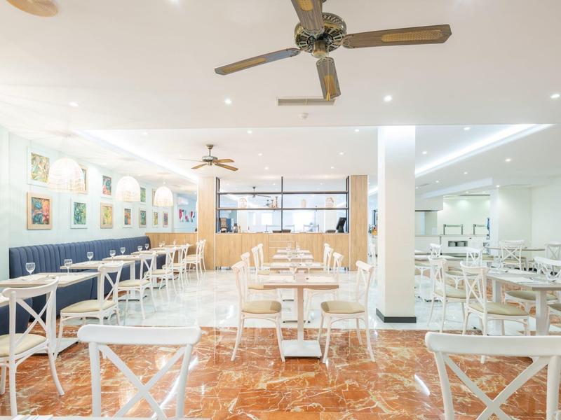 Helles, modernes Restaurant mit weißen Tischen und Stühlen, großem Fenster und Ventilatoren an der Decke.