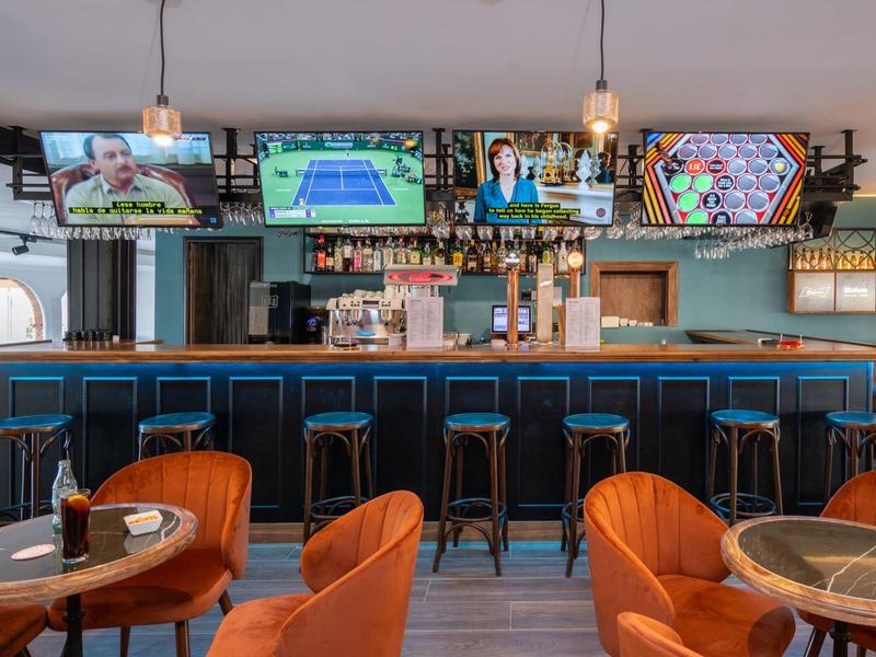 Moderne Bar mit blauer Theke, orangen Sesseln, hölzernen Tischen und mehreren Fernsehern an der Wand.