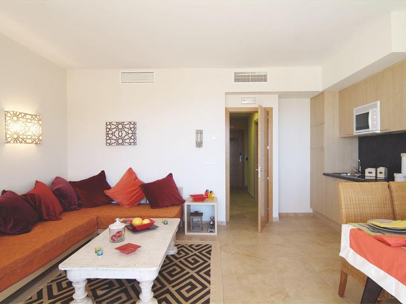 Helles Wohnzimmer mit orangem Sofa, weißen Wänden, Teppich und angrenzender Küche mit Bartresen.