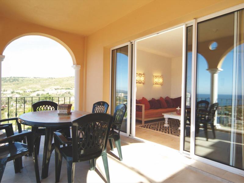 Terrasse mit runden Tisch und sechs schwarzen Stühlen, Blick auf grüne Landschaft und Meer.