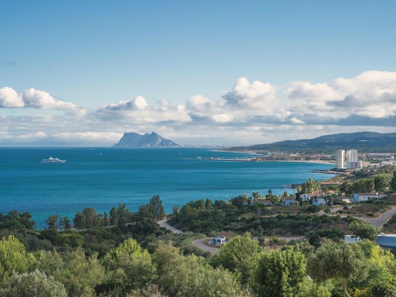 Küste mit blauem Meer, bewaldeten Hügeln, weißen Wolken und entfernten Bergen unter klarem Himmel.