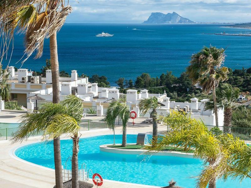 Blick auf blauen Pool mit Palmen, weißen Häusern und Meer mit Inseln im Hintergrund unter blauem Himmel.