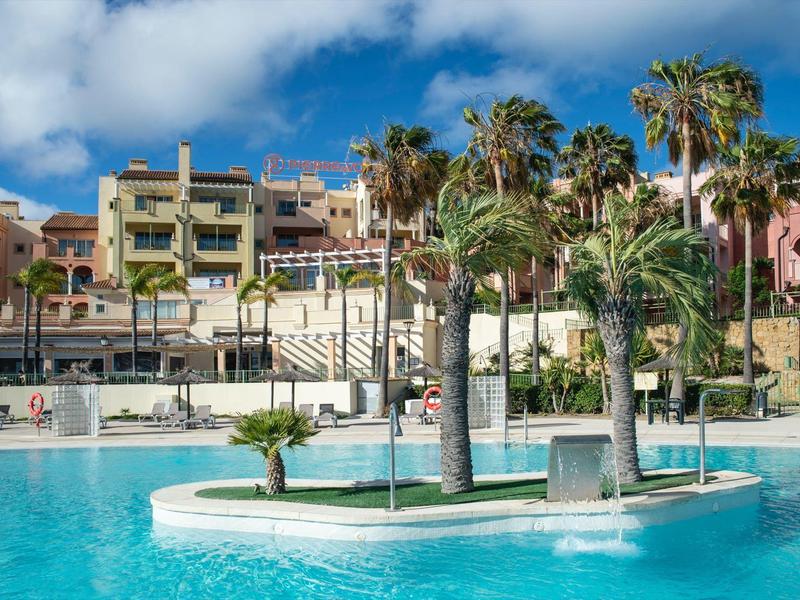 Großer blauer Pool mit Palmen und mediterranen Gebäuden im Hintergrund unter blauem Himmel.