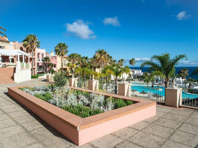 Blick auf sonnige Hotelanlage mit Pool, Palmen und Meer im Hintergrund unter blauem Himmel.