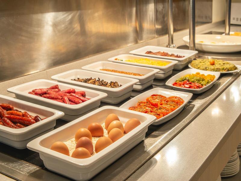 Buffet mit verschiedenen Speisen wie Brötchen, Wurst, Gemüse und Salat in weißen Behältern.