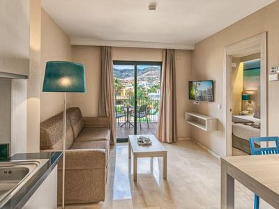 Helles Wohnzimmer mit Sofa, Tisch, TV, Küchenzeile und Balkon mit Blick auf grüne Berge.