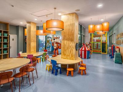 Moderner, bunter Kinderraum mit Tischen, Stühlen, Spielzeug und großen orangefarbenen Lampen.