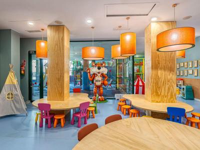 Bunter Spielraum mit Holztischen, Kindersesseln, Zelt und großen orangen Lampen an der Decke.