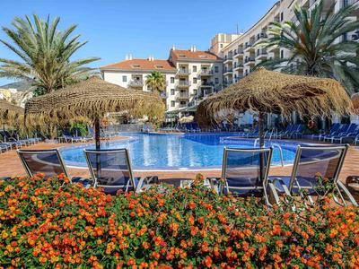 Schwimmbad mit Liegestühlen, Palmen und bunten Blumen vor einem mehrstöckigen Hotel.
