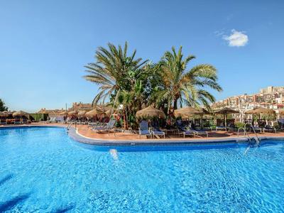 Großer Außenpool mit blauer Wasseroberfläche, Palmeninsel und zahlreichen Liegestühlen bei klarem Himmel.