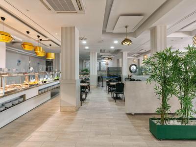 Hell eingerichteter Speiseraum mit Buffet, hohen Decken, Pflanzen und modernem Mobiliar.