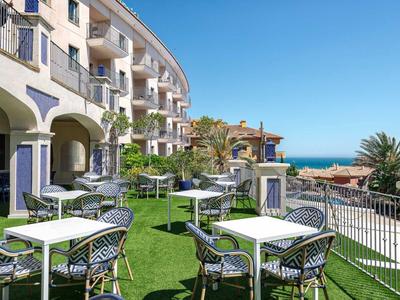 Moderner Außenbereich mit weißen Tischen, schwarzen Stühlen, grüner Rasenfläche und Meerblick.