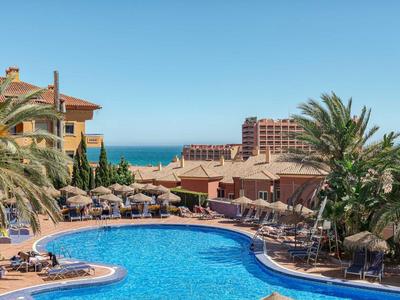 Großer, geschwungener Pool mit blauer Wasserfläche, umgeben von Sonnenschirmen und Palmen bei klarem Himmel.