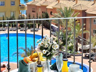 Balkontisch mit Frühstück, Blumen, Pool und Meer im Hintergrund an einem sonnigen Tag.