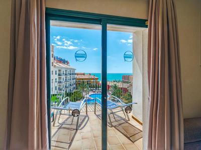 Blick durch Glasschiebetür auf Balkon mit Liegen, dahinter Meer und Häuser bei blauem Himmel.