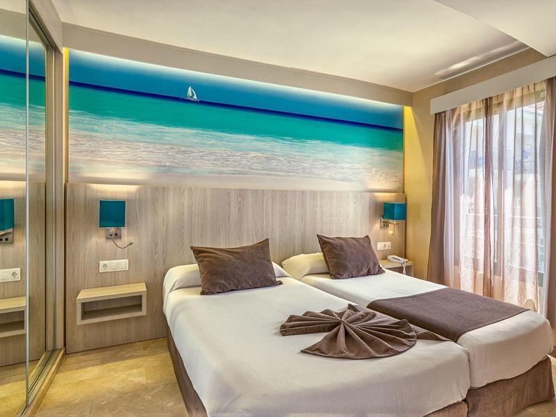 Modernes Schlafzimmer mit Doppelbett, Strandwandbild, beigen Vorhängen und zwei Nachttischen.