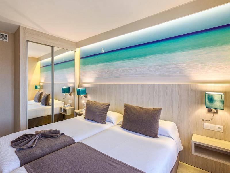 Modernes Hotelzimmer mit zwei Einzelbetten, grauen Kissen und Meereslandschaft als Kopfteilbeleuchtung.