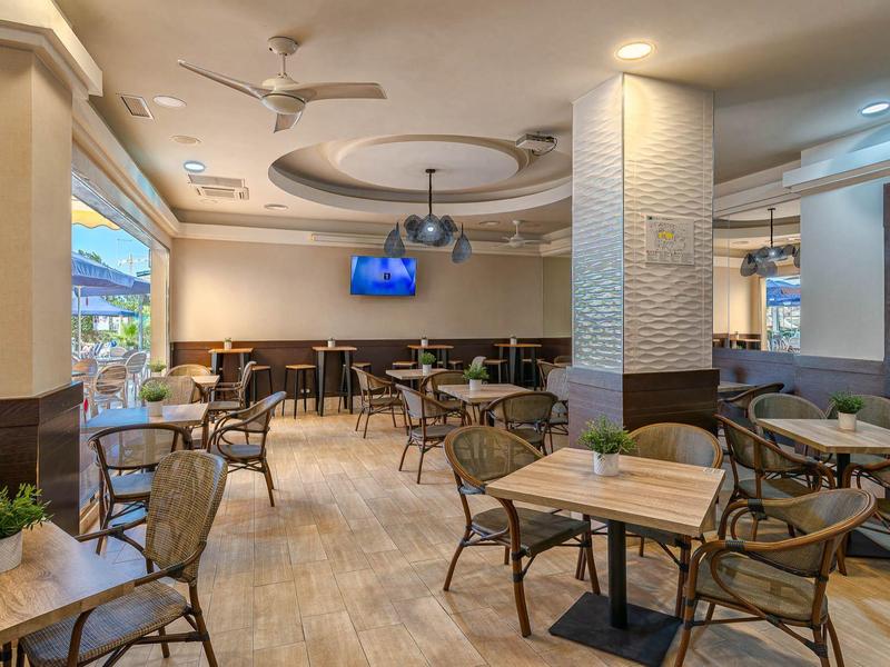 Modernes, helles Café mit Holzstühlen, kleinen Tischen und Deckenventilatoren, großer TV an der Wand.