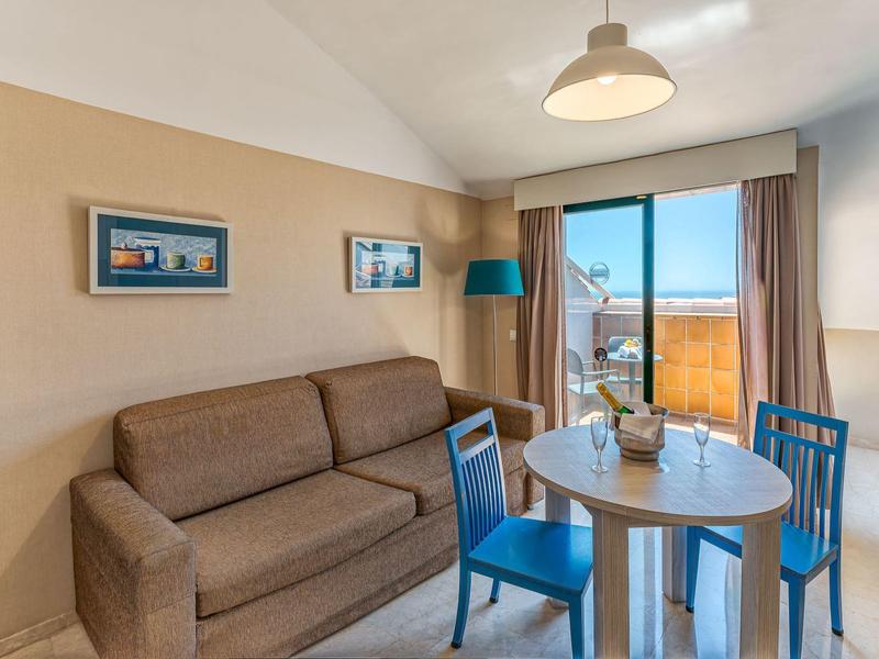 Wohnzimmer mit braunem Sofa, rundem Tisch, blauen Stühlen und Balkon mit Meerblick.