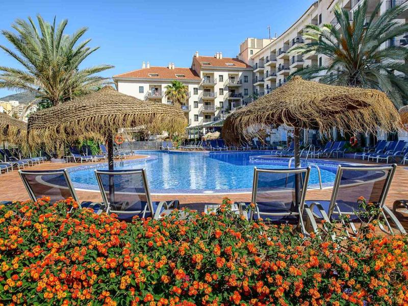 Schwimmbad mit Liegestühlen, Palmen und bunten Blumen vor einem mehrstöckigen Hotel.