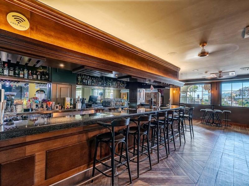 Leere Bar mit dunklem Holz, Theke, hohen Stühlen und großen Fenstern, sanftes Licht innen.