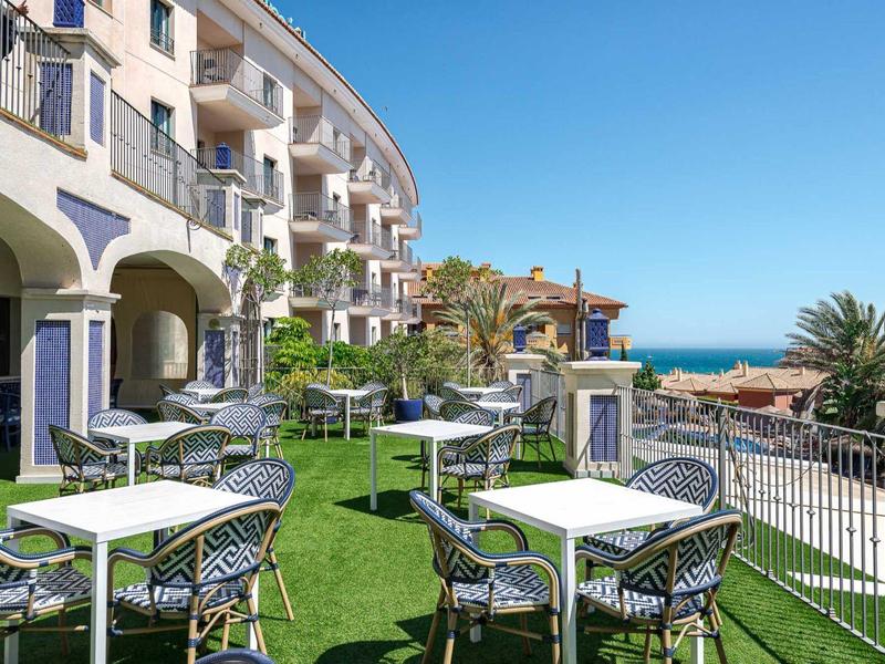 Moderner Außenbereich mit weißen Tischen, schwarzen Stühlen, grüner Rasenfläche und Meerblick.