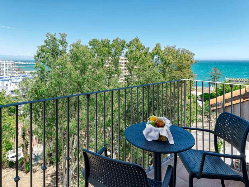 Balkon mit schwarzem Tisch, zwei Stühlen, Obstschale, Blick auf Bäume und Meer unter blauem Himmel.