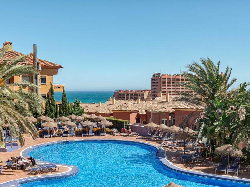 Großer, geschwungener Pool mit blauer Wasserfläche, umgeben von Sonnenschirmen und Palmen bei klarem Himmel.