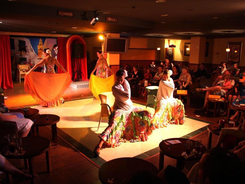 Theaterbühne mit vier Tänzern in leuchtenden Kostümen und Publikum in gedämpftem Licht.