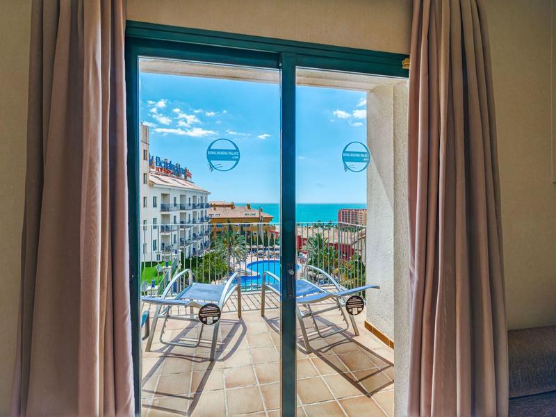 Blick durch Glasschiebetür auf Balkon mit Liegen, dahinter Meer und Häuser bei blauem Himmel.